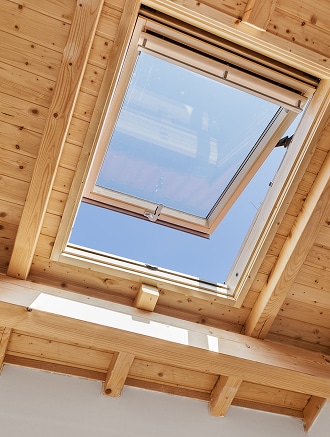 Fenêtre de toit mansarde en bois : lumière naturelle Puits de lumière de toit ouvrant en bois, laissant voir un ciel bleu clair, dans une structure au plafond à poutres et lambris apparents.