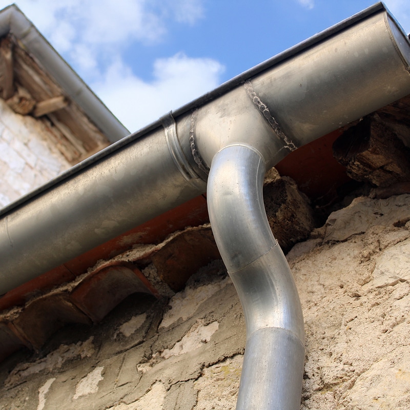Intérieur en cours de rénovation avec charpente en bois apparente, débris et planches sur un sol brut près d'une fenêtre baignée de lumière.