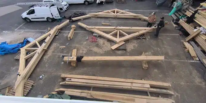 Assemblage charpente bois massif : ouvriers sur chantier Vue plongeante sur un chantier extérieur montrant l'assemblage de fermes en bois massif, deux ouvriers travaillent près des poutres.