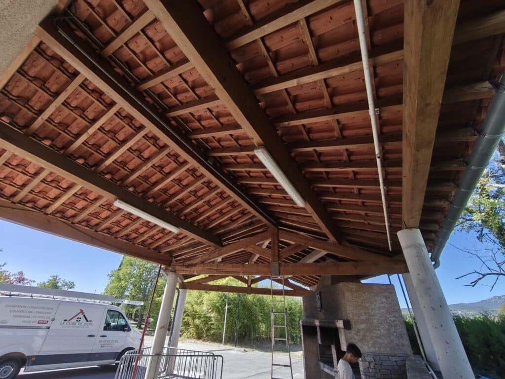 Auvent Bois et Tuiles : Finitions et Artisanat Vue sous un auvent avec charpente bois et tuiles TERREAL, présence d'un barbecue extérieur, d'un ouvrier, et d'une camionnette 'LE CUBE DE BOIS'.
