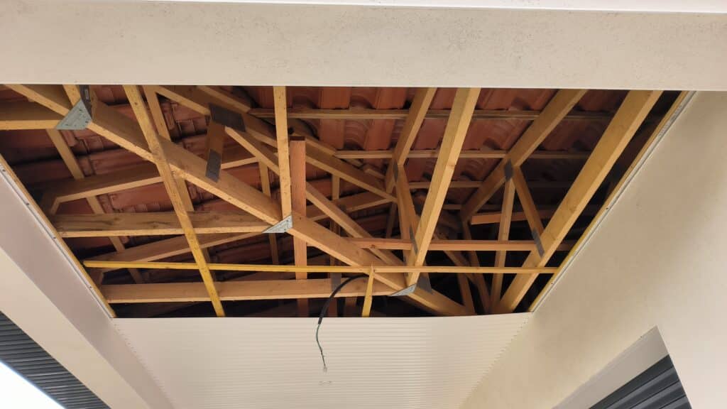 Charpente Bois Toit : Finition Plafond Terrasse Plafond de terrasse en cours de finition, laissant apparaître une structure de charpente en bois et des tuiles en terre cuite au-dessus. Un fil électrique pend.