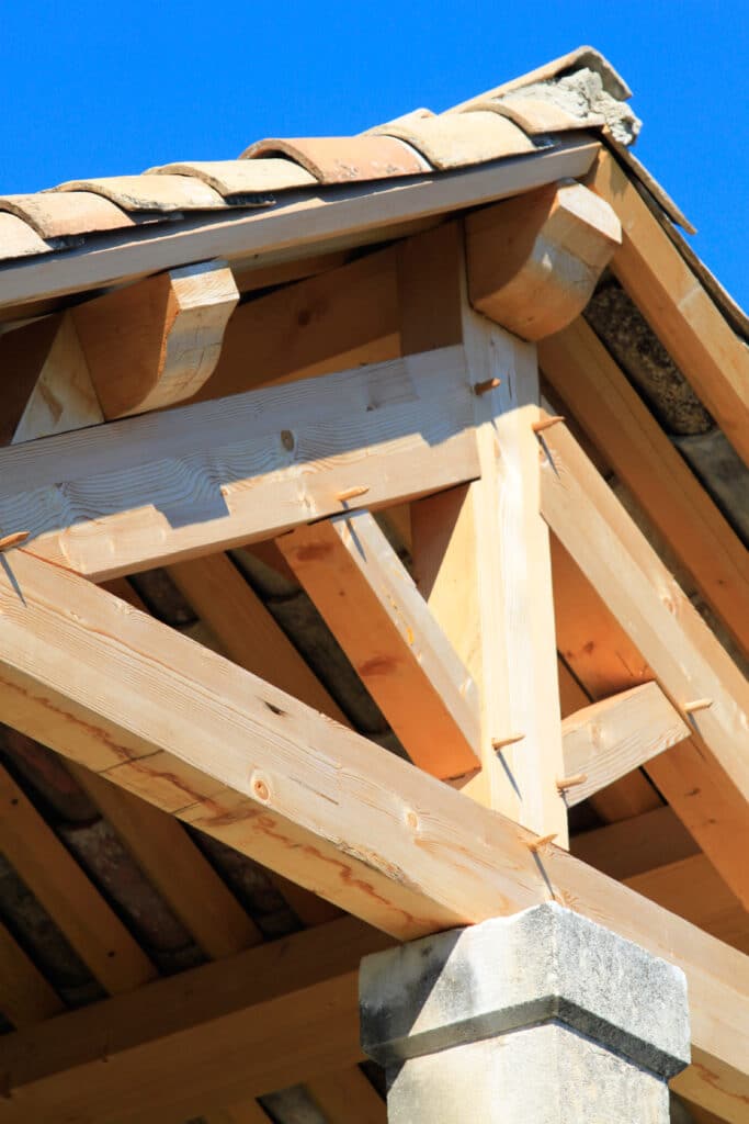 Charpente bois neuve, toiture tuiles canal, construction traditionnelle Charpente en bois clair neuve, avec chevrons et assemblages tenons-mortaises, reposant sur un pilier en pierre sous un ciel bleu et des tuiles canal.