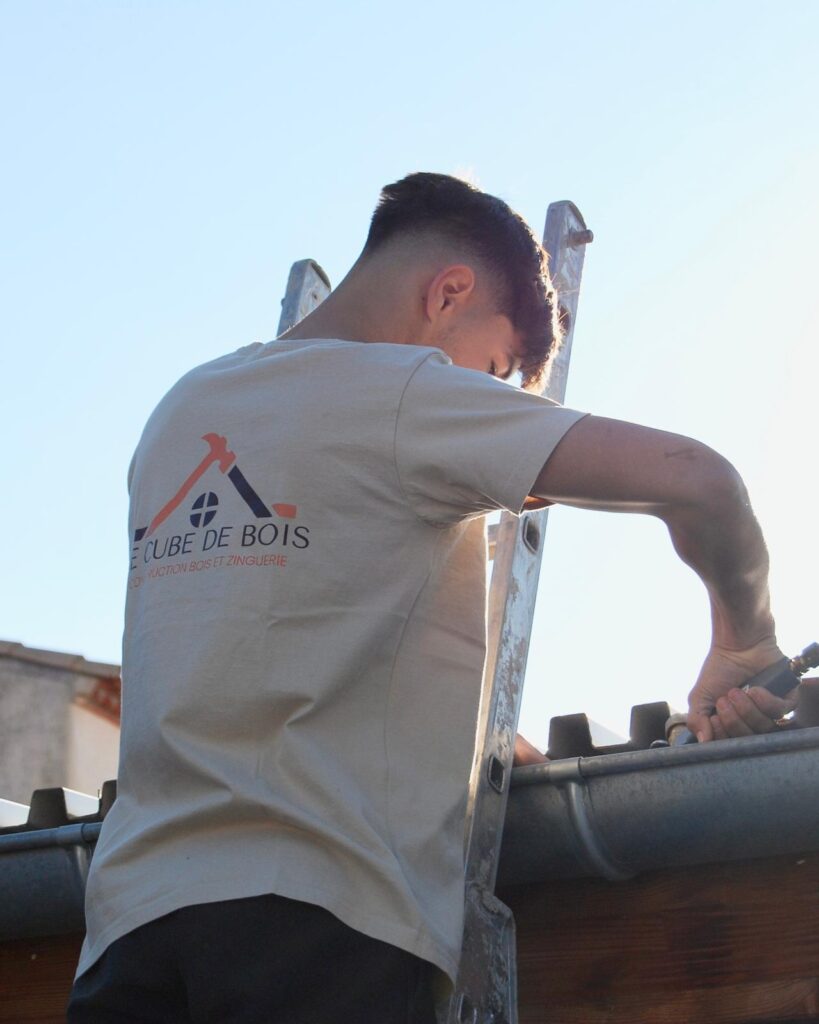 Couvreur Installant Gouttière - CUBE DE BOIS Jeune couvreur en t-shirt "CUBE DE BOIS CONSTRUCTION BOIS ET ZINGUERIE" montant une gouttière sur une échelle contre un ciel bleu lumineux.