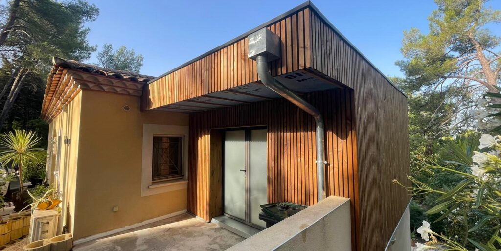 Extension Bardage Bois Moderne et Maison Traditionnelle Agrandissement moderne d'une maison, ajout de bardage bois vertical brun, avec gouttière métallique, terrasse et végétation luxuriante sous ciel bleu.