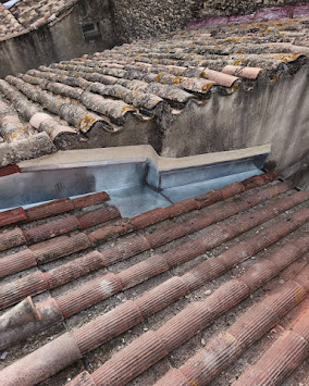 Détail de gouttière en zinc neuve réparant la jonction entre deux pans de toiture en tuiles anciennes et moussues, murs en maçonnerie.