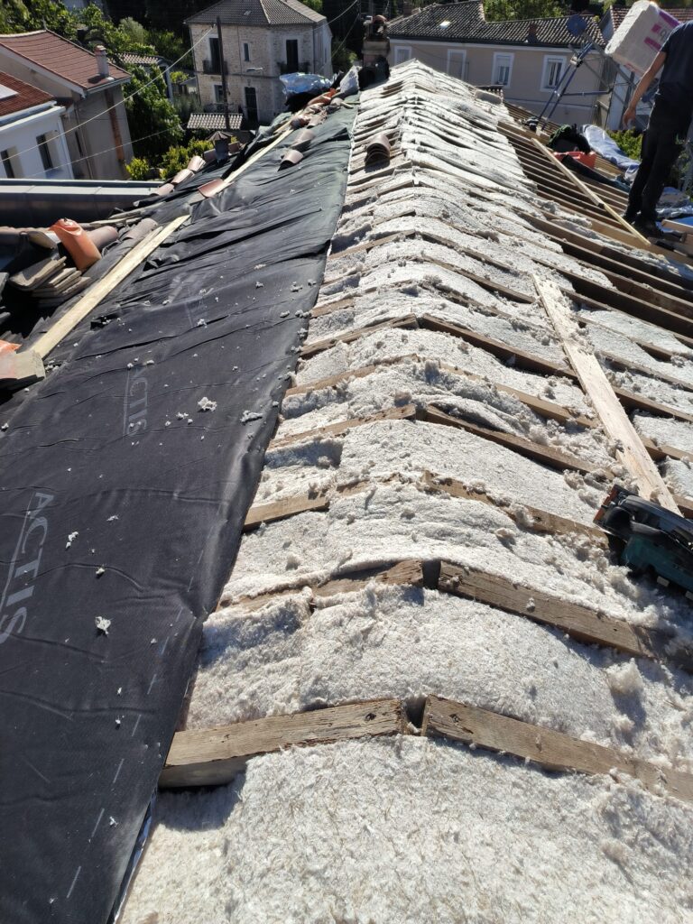 Isolation toiture : pose de laine isolante et membrane ACTIS Isolation de toiture avec laine isolante soufflée sous liteaux, membrane pare-vapeur noire ACTIS partiellement posée sur des tuiles retirées.