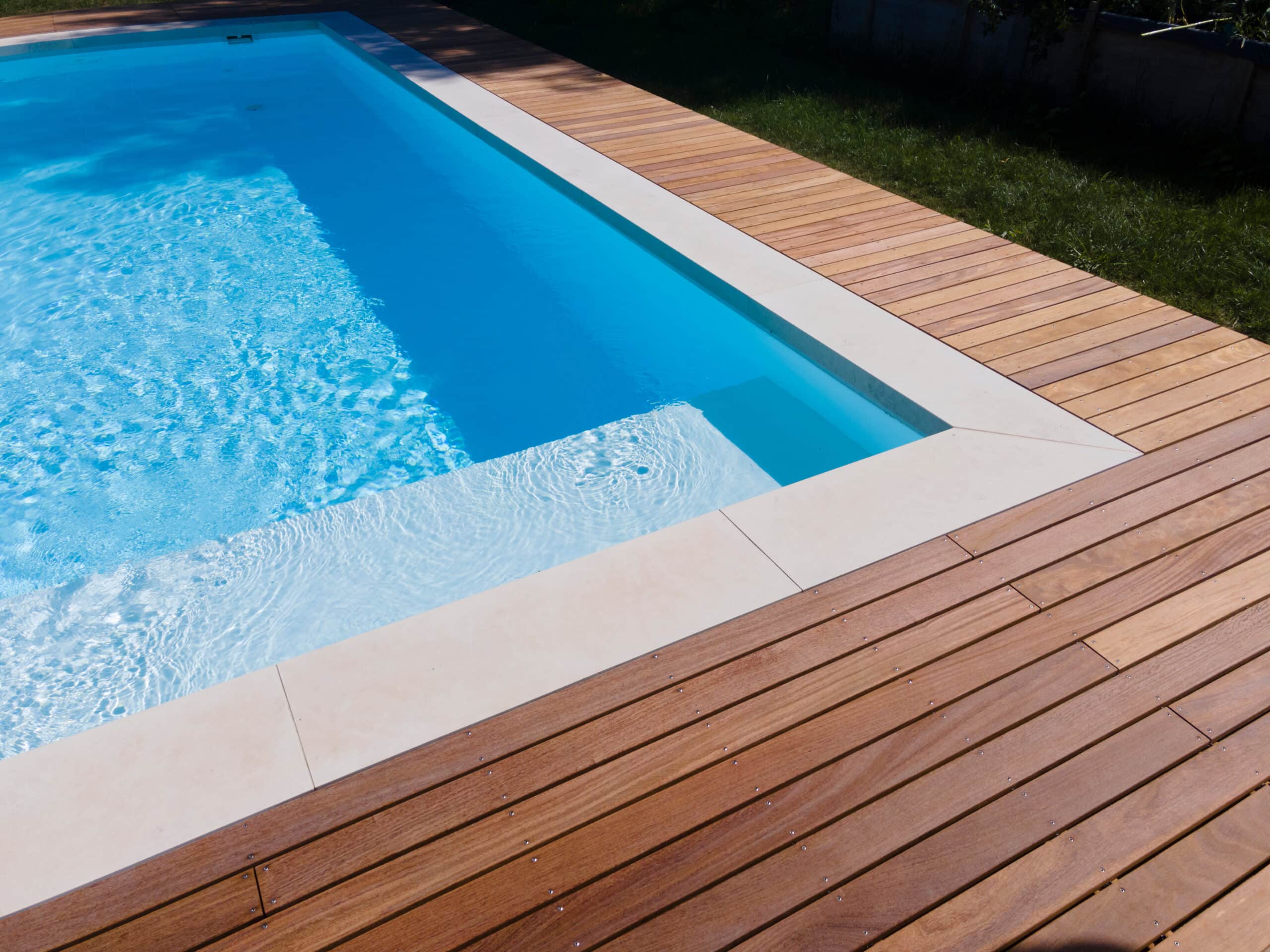 Angle d'une piscine rectangulaire moderne avec eau bleue cristalline et marches immergées, bordée d'une terrasse en bois brun clair.