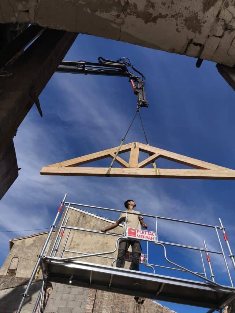 Pose de ferme de toit en bois par grue sur chantier Ouvrier sur échafaudage Plettac Mefran supervise la levée d'une ferme de toit en bois par une grue sous un ciel bleu.