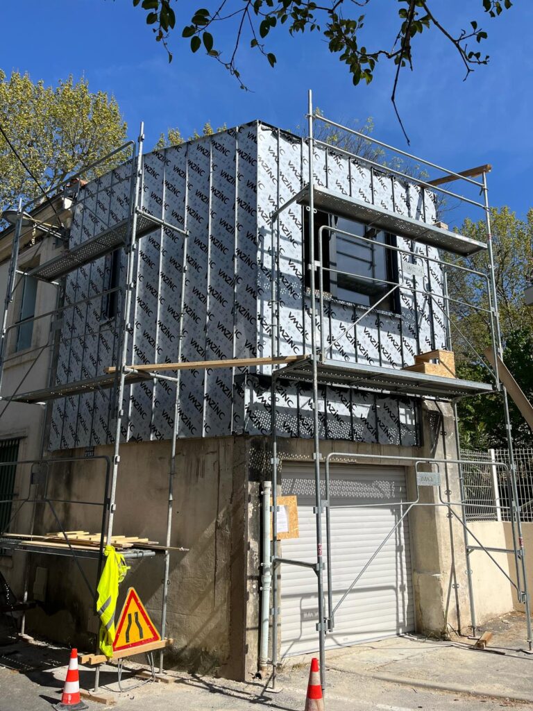 Rénovation façade : Isolation VMZINC et échafaudage Bâtiment en rénovation avec échafaudage, isolé par un matériau portant l'inscription 'VMZINC', signalisation de chantier et ciel bleu.