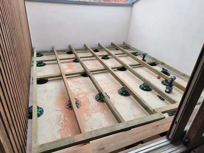 Pose Ossature Terrasse sur Plots Réglables : Bâtiment Construction d'une terrasse sur plots réglables noirs et verts, posant une ossature en bois traité sur une dalle de béton dans un espace extérieur avec bardage bois.