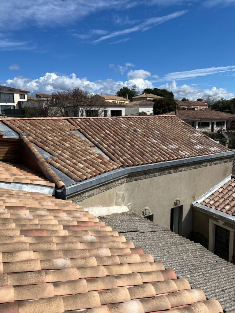 Toits en Tuiles Canal : Vue sur Quartier Résidentiel Vue plongeante sur des toits en tuiles canal rouges et grises sous un ciel bleu vif avec des nuages blancs, aperçu d'un quartier résidentiel.