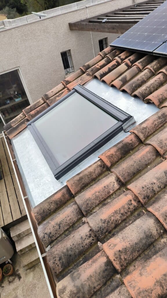 Installation Velux sur Tuiles Anciennes et Panneaux Solaires Fenêtre de toit installée sur un toit de tuiles romanes patinées, avec des panneaux solaires et une couverture métallique visible.